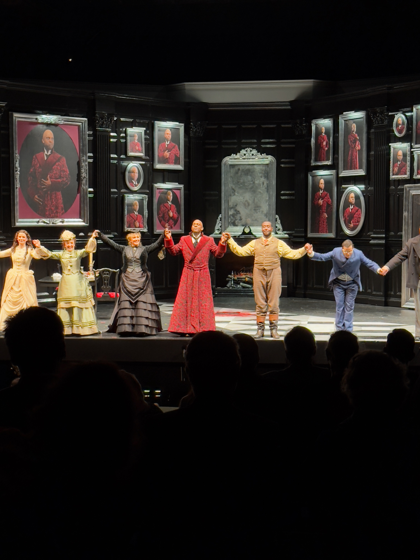 Full company of The Picture of Dorian Gray taking a bow on a black-paneled set hung with framed portraits of the title character in a red brocade robe