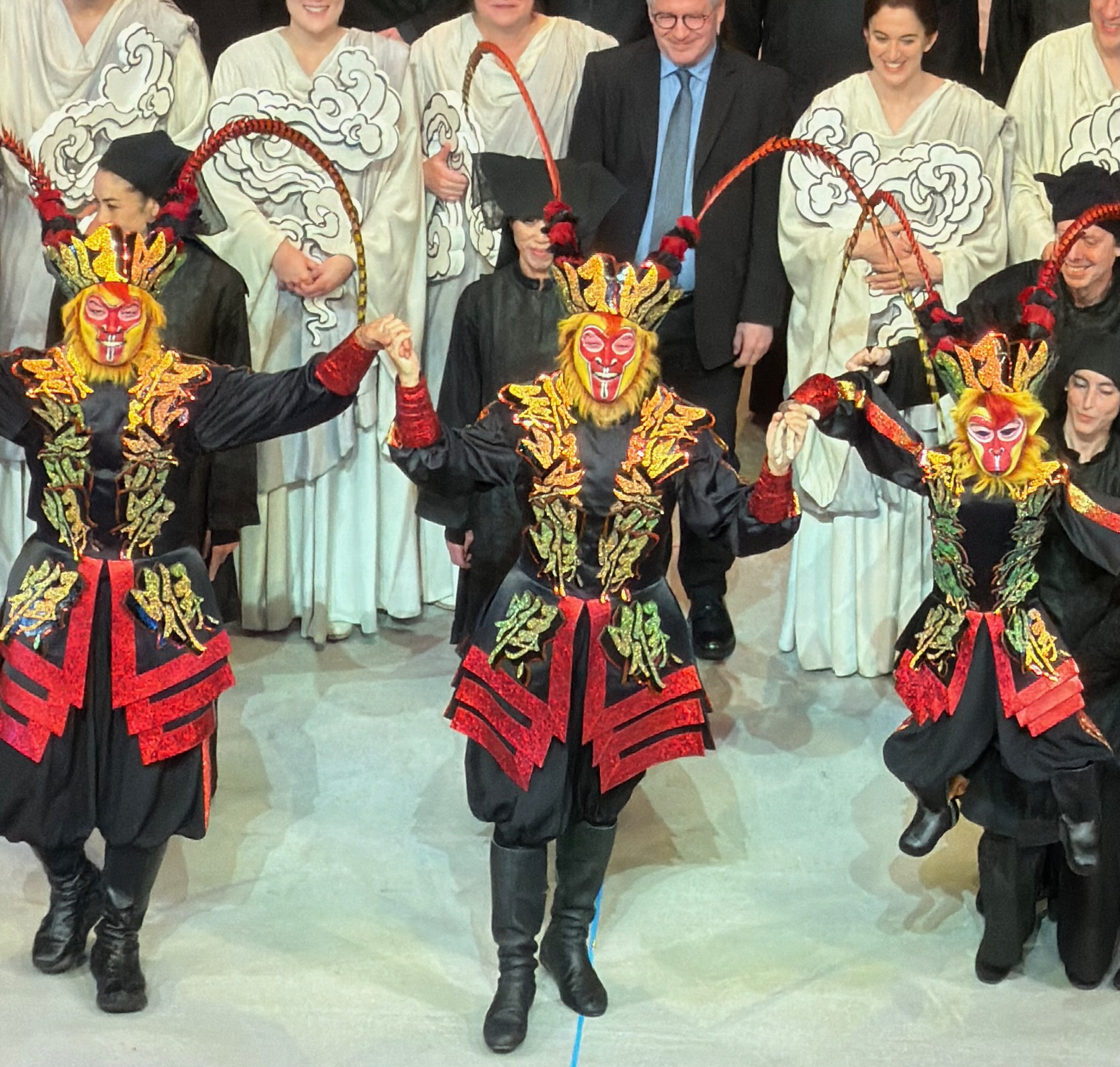Three performers in glittering red and gold monkey masks with long pheasant-feather plumes bow downstage, robed chorus in cloud-painted white behind